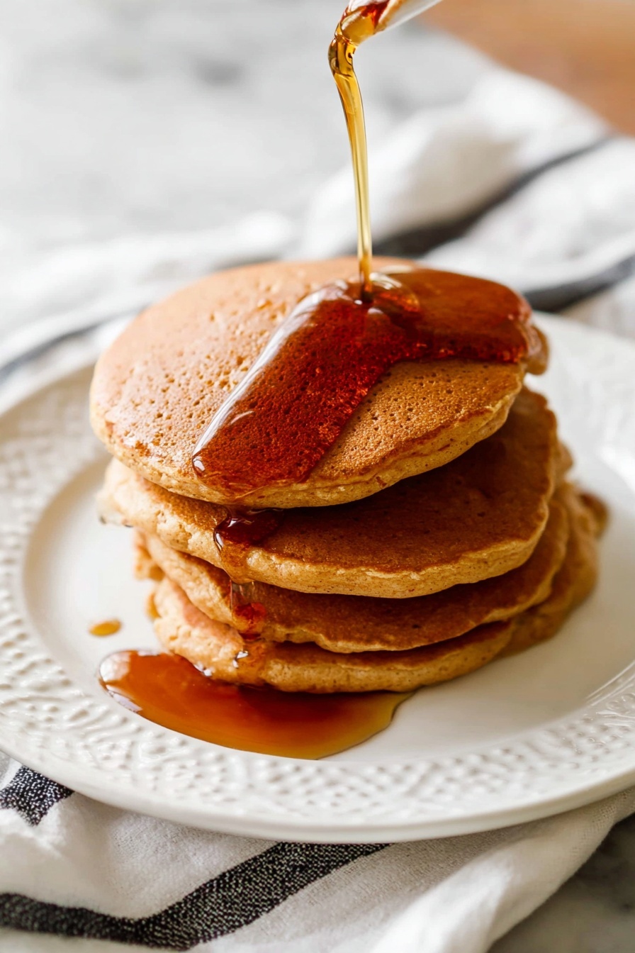 A stack of four light brown pancakes sits in the center of a white plate with a delicate, embossed pattern. The pancakes have a soft, fluffy texture with slightly darker edges. Dark amber syrup is being poured from above, slowly flowing down the top pancake and dripping over the sides, pooling slightly at the bottom edge on the plate. The plate rests on a white marbled surface with a white cloth featuring black stripes partly visible underneath. Photo taken with an iphone --ar 2:3 --v 7