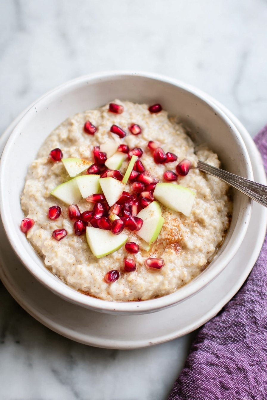 A white bowl filled with creamy oatmeal that has a light beige color and soft texture, topped with small bright red pomegranate seeds and thin light green apple slices scattered on top. There is a silver spoon resting inside the bowl on the right side, and the bowl sits on a white plate on a white marbled surface. A purple cloth is partially visible under the plate on the right side. Photo taken with an iphone --ar 2:3 --v 7